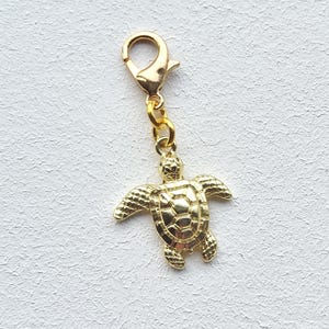 May include: A gold-toned metal charm in the shape of a sea turtle, featuring a detailed shell pattern and flippers. The turtle charm is attached to a small gold-toned lobster claw clasp by a circular jump ring, allowing for easy attachment.