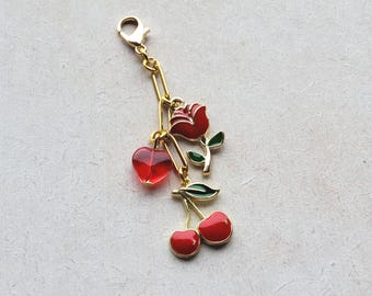 May include: Gold-coloured charm featuring red enamel heart, rose, and cherry pendants. The charm has a lobster clasp and chain link design. The rose has green leaves, and the cherries have gold accents.