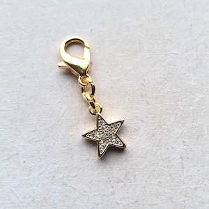May include: A gold-coloured charm featuring a star pendant. The star is adorned with small, clear crystals. The charm has a lobster clasp for easy attachment. The star charm is approximately 2 cm in size.