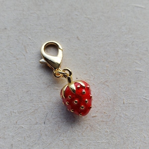 May include: A gold charm featuring a red enamel strawberry design. The charm has a lobster claw clasp.
