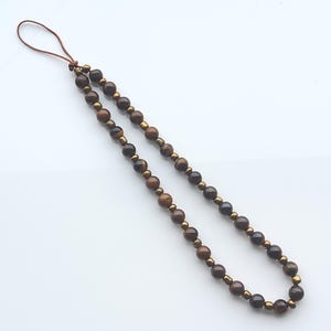 Puede incluir: Un collar de cuentas con un lazo de cordón de cuero marrón. El collar presenta una serie de cuentas marrones y doradas. Las cuentas están ensartadas para formar un lazo, adecuado para usar o como accesorio decorativo.