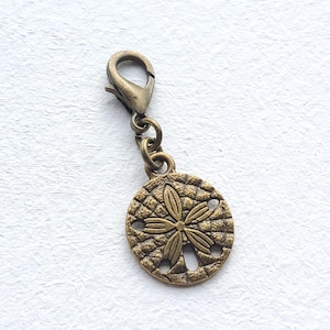May include: A bronze-coloured charm featuring a sand dollar design. The round charm has a detailed floral pattern in the centre and small holes around the edges. It is attached to a lobster clasp, suitable for a necklace or bracelet.