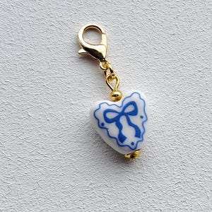 May include: A heart-shaped charm featuring a white ceramic base with a blue painted bow design. The charm is attached to a gold-toned metal lobster clasp by a small gold-toned jump ring and a gold-toned bead spacer at the top and bottom.