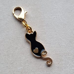 Op de afbeelding: Een goudkleurige kattenbedel met een zwarte body en een gouden hartdetail. De bedel heeft een goudkleurige sluiting en een gekrulde staart. Een sieradenaccessoire.