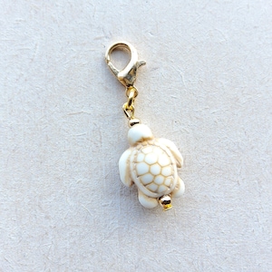 May include: A white and tan turtle charm with a gold-colored clasp and chain. The turtle has a detailed shell pattern and a small gold bead at the bottom. This charm is a decorative accessory.