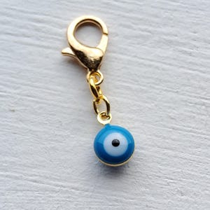 May include: A gold-coloured charm featuring a lobster clasp and a small blue eye pendant. The pendant has a white iris and a black pupil, with a gold-coloured border. The charm is designed to be attached to a bracelet or necklace.