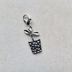 May include: Silver charm featuring a small potted plant with a textured pot and a simple stem and leaves.