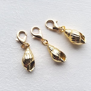 May include: Three gold-coloured seashell charms with lobster claw clasps. The charms are detailed with ridges and curves, and are displayed on a white surface. These charms are suitable for jewellery making or other craft projects.