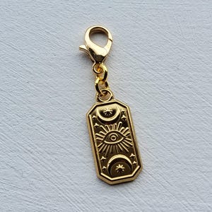Könnte beinhalten: Ein goldfarbener Anhänger mit Karabinerverschluss. Der rechteckige Anhänger zeigt ein eingraviertes Design eines Auges, Sonnenstrahlen, Monde und Sterne. Der Anhänger wird wahrscheinlich für Schmuck oder Accessoires verwendet.