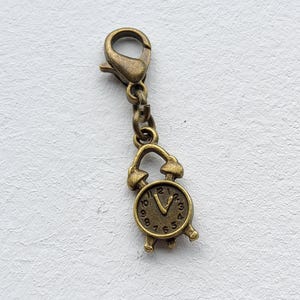 May include: A bronze-colored charm featuring a small clock design. The clock has a detailed face with numbers and hands, and is attached to a clasp and chain. The charm is likely used for jewelry or crafts.