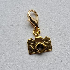 May include: A gold-coloured camera charm with a lobster clasp. The charm is a miniature replica of a vintage camera, featuring detailed design elements. The camera charm is suspended from a gold-toned clasp, suitable for jewellery or craft projects.