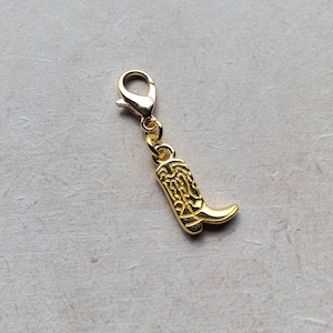 May include: A gold-coloured charm featuring a detailed cowboy boot design. The boot charm is attached to a lobster clasp, allowing it to be easily added to a bracelet or necklace. The charm is made of a shiny, metallic material.