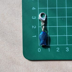 May include: A blue fish charm with a silver lobster clasp. The charm is hanging from a green cutting mat with numbered grid lines.