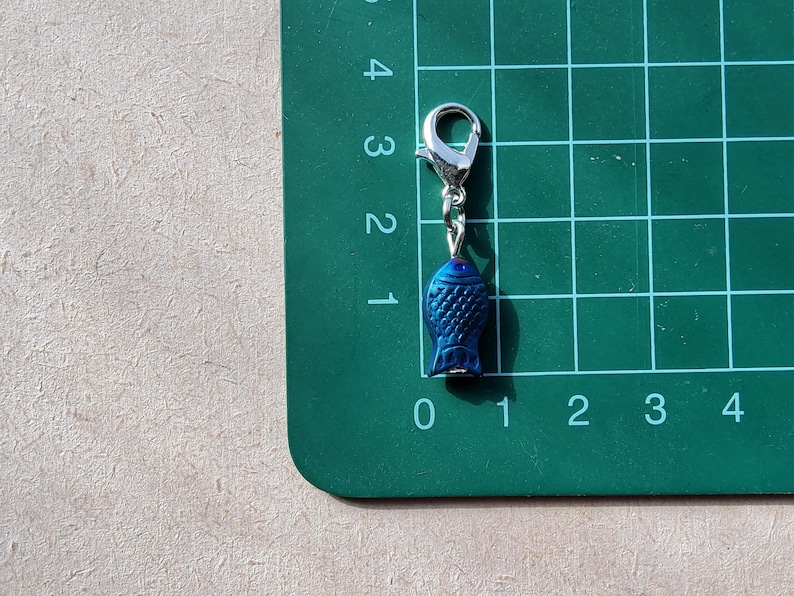 May include: A blue fish charm with a silver lobster clasp. The charm is hanging from a green cutting mat with numbered grid lines.