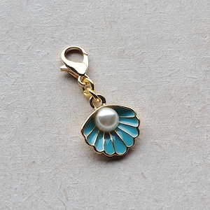 May include: A gold-colored charm with a blue seashell design and a white pearl in the center. The charm has a gold loop for attaching to a keychain or bag.