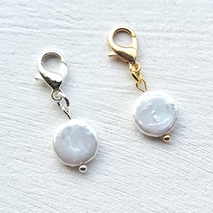 Könnte beinhalten: Zwei Anhänger mit weißen, münzförmigen Perlen. Einer hat einen silberfarbenen Verschluss und Akzente, der andere goldfarbene Beschläge. Beide haben eine kleine, runde Akzentperle am unteren Ende der Perle.