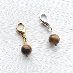 Op de afbeelding: Twee bedelhangers met tijgeroogkralen. Eén hanger heeft een goudkleurige sluiting en ketting, de andere een zilverkleurige sluiting en ketting. De ronde kralen hebben bruine en gouden patronen.