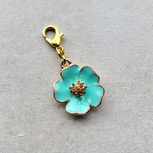 Puede incluir: Un charm dorado con una flor de esmalte azul claro. La flor tiene un centro dorado con un diseño detallado.