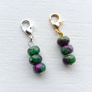 May include: Two charm pendants with lobster clasps. One has a silver-tone clasp, the other gold-tone. Each pendant features three beads of green and purple stone. The beads are stacked and connected to the clasp with a small metal loop.