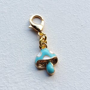 Könnte beinhalten: Ein goldfarbener Charm mit einem Pilzdesign. Der Pilz hat eine hellblaue Kappe mit weißen Punkten und einen türkisfarbenen Stiel. Der Charm ist an einem goldfarbenen Verschluss und einer Kette befestigt.