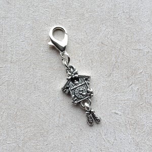 Könnte beinhalten: Ein silberfarbener Charm-Anhänger mit einem Miniatur-Kuckucksuhr-Design. Der Anhänger hat einen Karabinerverschluss zur einfachen Befestigung an einem Armband oder einer Halskette. Das Zifferblatt ist sichtbar, und der Anhänger hat herabhängende Gewichte.