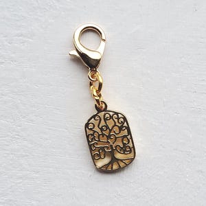 May include: A gold-coloured charm featuring a tree of life design. The charm has a rectangular shape with a cutout tree pattern and a gold-coloured clasp. It is attached to a gold-coloured chain.