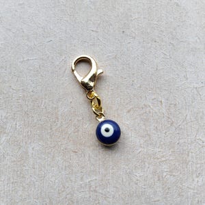 Pode incluir: Um berloque dourado com um fecho lagosta e uma pequena conta redonda azul e branca de olho grego. O berloque foi projetado para ser preso a uma pulseira, colar ou outra joia.