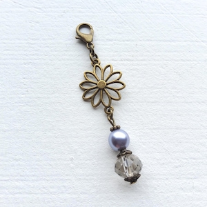 Op de afbeelding: Een decoratieve bedel met een bronzen bloemvormige hanger, een lavendelkleurige parel en een facetgeslepen grijze kristallen kraal. De bedel heeft een karabijnsluiting. Het ontwerp is delicaat en vrouwelijk.