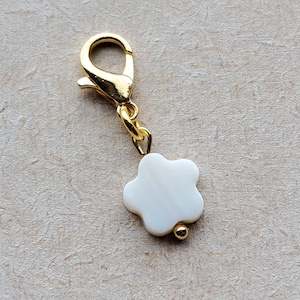 Könnte beinhalten: Ein goldfarbener Karabinerverschluss-Anhänger mit einem weißen, blütenförmigen Anhänger und einer kleinen goldenen Perle. Der Anhänger ist für die Schmuckherstellung oder als Accessoire gedacht.