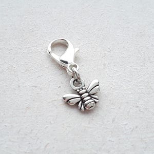 May include: A silver-toned metal charm in the shape of a bee, featuring detailed wings and a segmented body. The bee charm is attached to a functional silver-toned lobster claw clasp, designed for easy attachment to bracelets, necklaces, or other items.