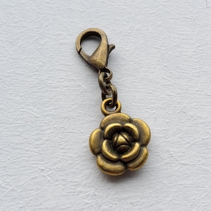 May include: A bronze-coloured charm featuring a rose flower design. The charm is connected to a lobster clasp with a small chain. The rose has detailed petals and a vintage aesthetic. Suitable for jewellery making or keychains.