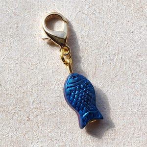May include: A gold-toned metal charm with a blue fish design. The fish has a textured surface and a gold-toned loop at the top.