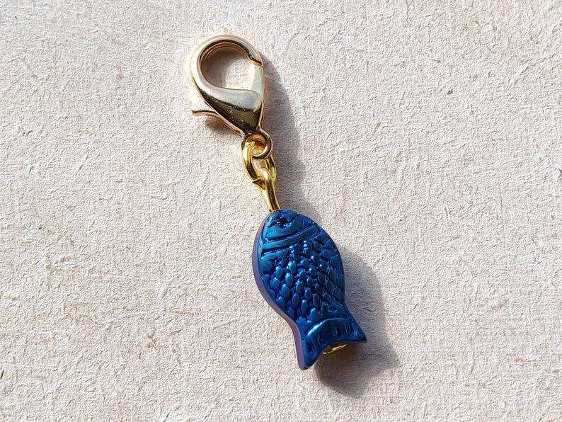 May include: A gold-toned metal charm with a blue fish design. The fish has a textured surface and a gold-toned loop at the top.