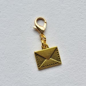 May include: A gold-coloured charm depicting a small envelope. The charm is connected to a gold-coloured lobster clasp. The envelope has a detailed design with a flap and dotted edges. The charm is likely for a bracelet or necklace.