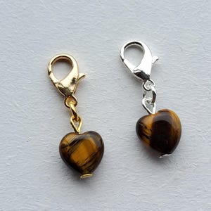 Op de afbeelding: Twee hartvormige bedels met karabijnsluitingen. De ene bedel heeft een goudkleurige sluiting en ketting, de andere een zilverkleurige sluiting en ketting. Beide hebben een gepolijste tijgeroogsteen in hartvorm.