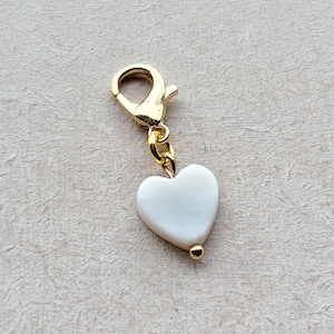 Könnte beinhalten: Ein goldfarbener Charm mit einem weißen herzförmigen Anhänger. Der Charm hat einen Karabinerverschluss und eine kleine goldene Perle am unteren Ende des Herzens. Der Herzanhänger ist glatt und hat einen perlmuttartigen Glanz.