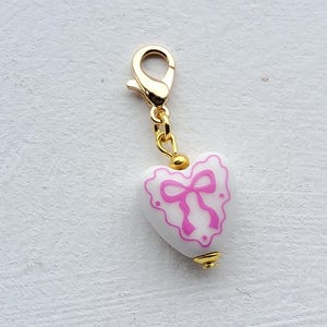 May include: A white heart-shaped charm with a pink bow design and gold-colored accents. The charm is attached to a gold-colored clasp, suitable for a bracelet or necklace.