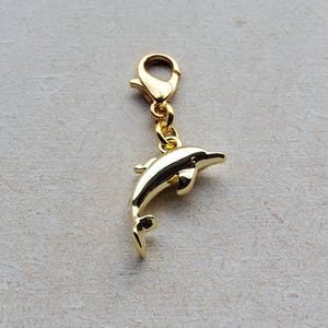 May include: A gold dolphin charm with a lobster clasp. The charm is a three-dimensional rendering of a dolphin, with details such as fins and a curved body. The charm is designed to be attached to a bracelet or necklace.