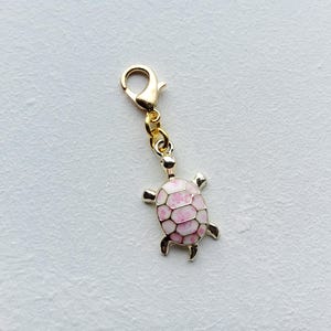 Op de afbeelding: Een goudkleurige schildpadbedel met een karabijnsluiting. De schildpad heeft een roze en witte schild en zilverkleurige poten en kop. De bedel is waarschijnlijk gemaakt van metaal en is ontworpen om aan een armband of ketting te worden bevestigd.