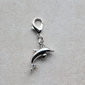 May include: A silver dolphin charm with a lobster clasp. The dolphin has detailed fins and a curved body, and is attached to the clasp by a small chain. The charm is likely made of metal and is designed to be attached to a bracelet or necklace.