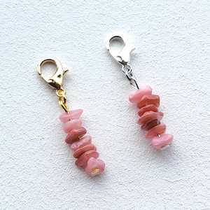 Puede incluir: Dos dijes colgantes con fragmentos de piedra pulida en tonos rosa y marrón rojizo ensartados verticalmente. Un dije tiene un cierre de langosta en tono dorado, mientras que el otro presenta un cierre de langosta en tono plateado.