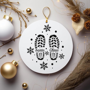 Puede incluir: Un adorno navideño blanco con un colgador dorado. El adorno presenta huellas negras con las palabras "Santa Claus" y diseños de copos de nieve. Adornos y elementos decorativos navideños dorados y blancos rodean el adorno.