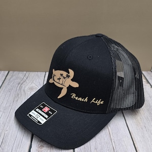 May include: Black trucker hat with a mesh back and a curved brim. The hat features a brown leather patch with a turtle and palm tree design, and the words "Beach Life" in gold script. The hat is adjustable.