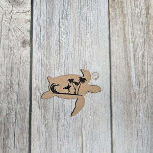 Könnte beinhalten: Hellbraunes Holzschild in Schildkrötenform mit ausgeschnittener Strandszene. Die Szene umfasst eine Welle, einen Surfer und Palmen. Das Ornament hat ein kleines Loch zum Aufhängen.