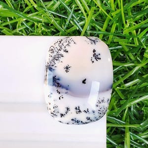 May include: A smooth, oval-shaped dendritic agate cabochon featuring a translucent, milky white base with intricate, dark black fern-like inclusions scattered across the surface, displaying a polished finish and a rounded, domed profile.
