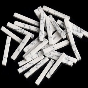 Può includere: Una collezione di perline rettangolari in pietra bianca con un motivo marmorizzato nero e grigio. Le perline sono sparse su una superficie nera, evidenziando la loro finitura lucida e le variazioni naturali. Ideale per la creazione di gioielli.