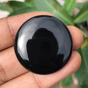 Könnte beinhalten: Ein glatter, kreisförmiger, polierter schwarzer Edelstein-Cabochon mit Hochglanz-Finish. Die Oberfläche ist undurchsichtig und reflektierend, mit einem hellen weißen Lichtreflex auf der linken Seite, der die abgerundete, gewölbte Form betont.