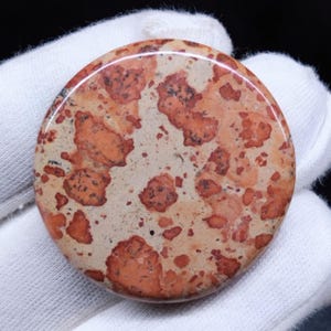 Puede incluir: Una piedra redonda y pulida con un patrón moteado. La piedra presenta un color base beige con manchas rojizas-anaranjadas y marrón oscuro. La superficie es lisa y brillante, con un aspecto natural y terroso.