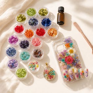 Puede incluir: Un surtido de flores secas de colores en pequeños recipientes transparentes, una botella marrón, una funda transparente para teléfono, un colgante y arte de uñas, todos con diseños florales. Las flores son en tonos de rojo, naranja, morado, azul y verde.