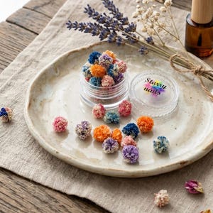 Puede incluir: Un tarro transparente lleno de pequeños capullos de flores secas de colores, con el texto "Starry nails" en la tapa. Los capullos son de color rosa, naranja, azul y morado. El tarro está sobre un plato de cerámica con lavanda seca.